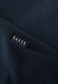 Tumšs zili audums ar strukturētu pinumu, kura papildinājums ir redzama birka ar tekstu "BAKER by TED BAKER" baltajās burtiņās.