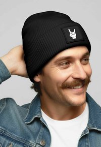Black ribbed beanie with a rectangular patch featuring a white hand gesture. Soft texture and fitted design. No additional embellishments.