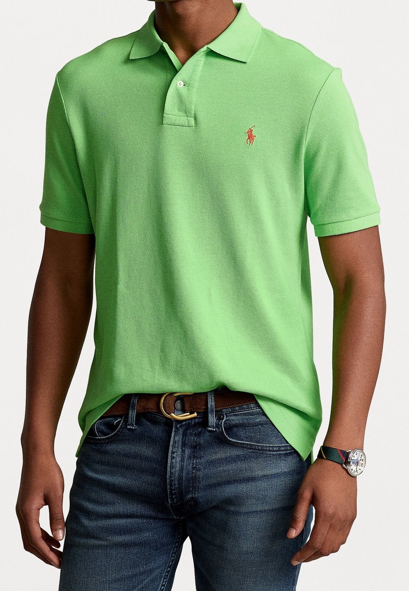Homme portant une chemise polo vert clair rentrée dans un jean bleu foncé, avec une ceinture marron et une montre-bracelet avec un bracelet à rayures.