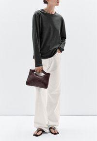 Maglietta a maniche lunghe grigio scuro, pantaloni bianchi a gamba larga e una borsa intrecciata bordeaux con manico, tenuta in una mano. Infradito neri.