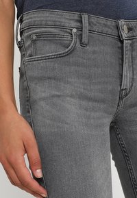 Jeans de mezclilla gris con un diseño ajustado, cinco bolsillos y detalles de costura en contraste a lo largo de las costuras y la cinturilla.