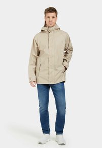 Beige wasserdichte Jacke mit Kapuze, Frontreißverschluss, seitlichen Taschen und elastischen Bündchen, kombiniert mit blauen Jeans und weißen Turnschuhen.