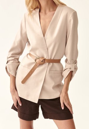 Beiger Wickelblazer mit hochgekrempelten Dreiviertelärmeln und beigem Leder Gürtel, kombiniert mit dunkelbraunen, umgeschlagenen Shorts.