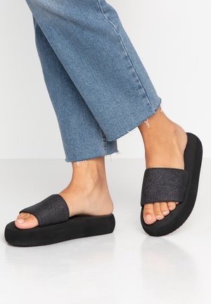 Füße in schwarzen Plateausandalen und blauen, gekürzten Jeans vor einem schlichten hellen Hintergrund.