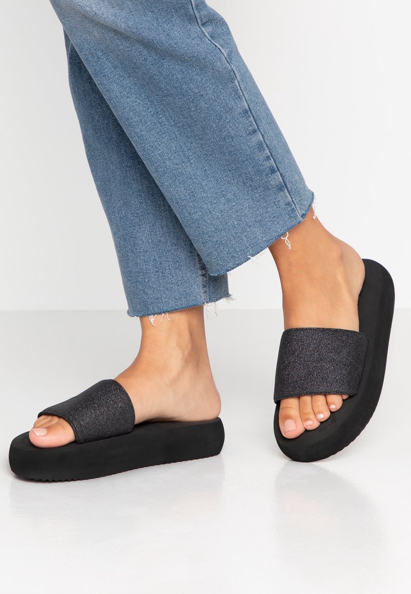 Pieds en sandales à plateforme noires et jean bleu court sur un fond clair uni.