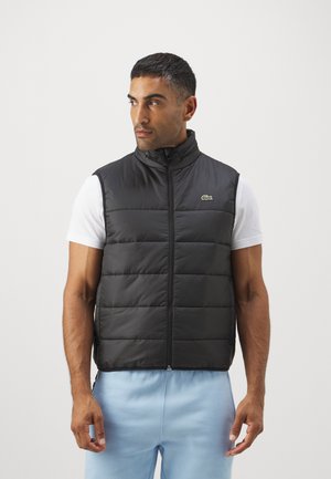 Zwarte gewatteerde gilet met hoge kraag en mouwloos ontwerp. Voorzien van een voorzijde rits en een klein groen logo op de borst.