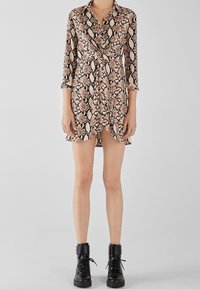 Abito camicia a fantasia di pelle di serpente in beige e nero, con cintura in vita, colletto e maniche lunghe. Abbinato a stivaletti neri alla caviglia.