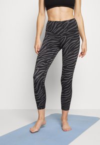 Frau, die barfuß auf einer blauen Yogamatte steht und schwarze und graue, zebra-gestreifte Leggings sowie einen schwarzen Sport-BH trägt.