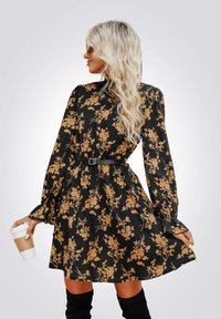 Abito floreale nero con maniche lunghe e vita cintrata, decorato con motivi di fiori in tono oro e una cintura nera. Il modello tiene una tazza di caffè.