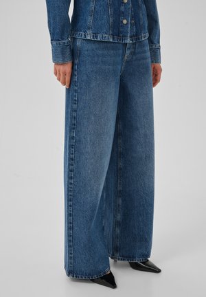 Personne portant un jean en denim bleu à jambes larges et une veste en denim boutonnée assortie, associée à des chaussures noires à bout pointu.