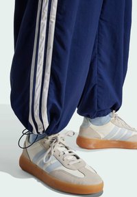 Marineblaue Trainingshosen mit drei weißen Streifen; graue und hellblaue Sneakers mit Gummisohle; Merkmale sind elastische Bündchen und Schnürsenkel.