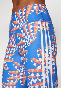 Leggings azuis com um padrão geométrico de triângulos em vermelho, branco e amarelo. Inclui três riscas brancas de lado.