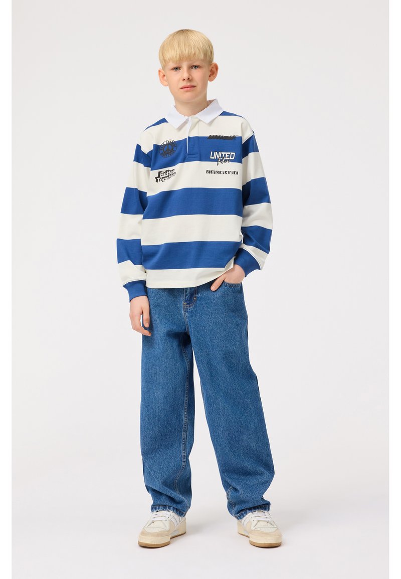 Camiseta de rugby a rayas en azul y blanco, con cuello y detalles del logo, combinada con jeans de denim azul de pernera ancha y zapatillas beige.