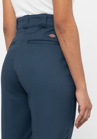 Midjeförsedda mörkblå byxor i mjukt tyg, med platt framstycke, sydda fickor och en liten logotyp på bakre midjeresåren.