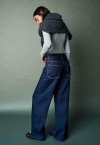 Ženska s temnimi lasmi, oblečena v sivo majico z dolgimi rokavi, temne široke kavbojke, temne čevlje in oglast pulover, ki ji visi čez ramena.