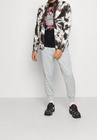 Svart och vit tie-dye jacka över en svart grafisk t-shirt, kombinerat med grå sweatpants och svarta sneakers med röda accenter.