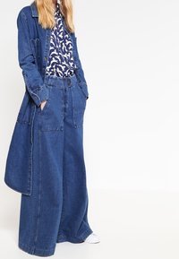 Giacca di denim con tasche a toppa sopra una camicia blu a fantasia, abbinata a pantaloni di denim a gamba larga. Tutti i pezzi sono di diverse tonalità di blu.