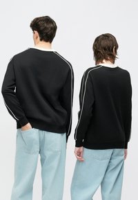 Fekete pulóverek fehér oldalsávokkal, világoskék laza fazonú farmernadrággal párosítva, hátulnézetből.