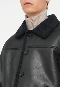 Schwarze Lederjacke mit strukturierter Oberfläche, ausgestattet mit einem flauschigen schwarzen Kragen, zwei Knöpfen und einem beigen gerippten Rollkragenpullover darunter.