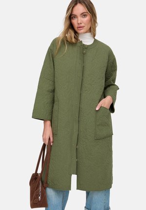 Junge Frau mit langem, gestepptem olivgrünem Mantel, hellblauen Jeans, weißem Rollkragenpullover, die eine braune Veloursledertasche hält.