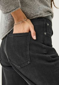 Jeans in denim nero con taglio dritto, dotati di tasche posteriori, cuciture discrete e un maglione in maglia grigia. Dettaglio della mano riposta nella tasca.