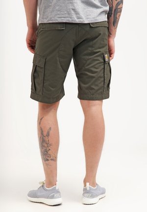 Person trägt olivgrüne Cargoshorts mit Seitentaschen, graue Nike-Sneaker und hat ein detailliertes Kaninchen-Tattoo an der linken Wade, steht vor einem weißen Hintergrund.