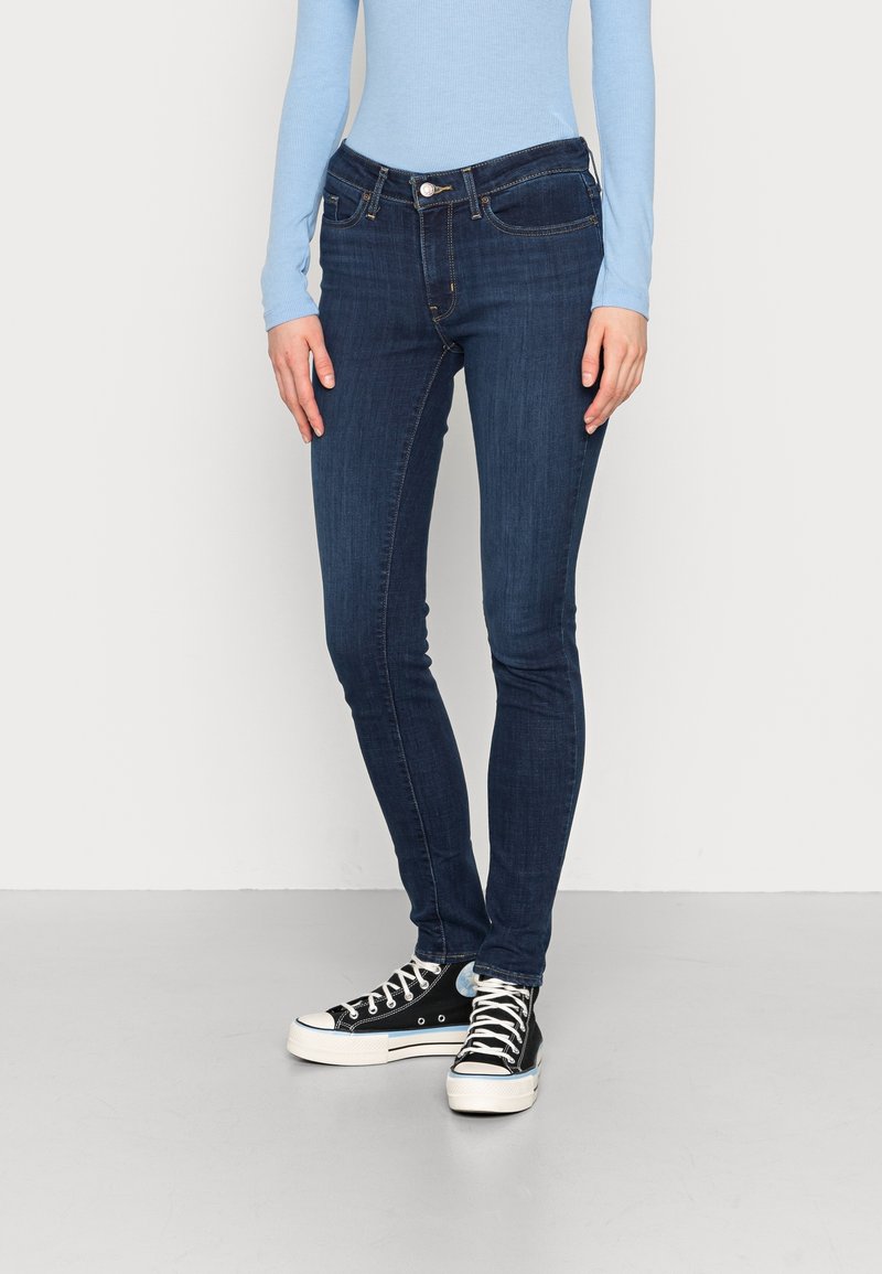 Jean skinny bleu foncé avec des coutures, cinq poches et une fermeture à bouton, porté avec des baskets montantes noires et un haut bleu clair.