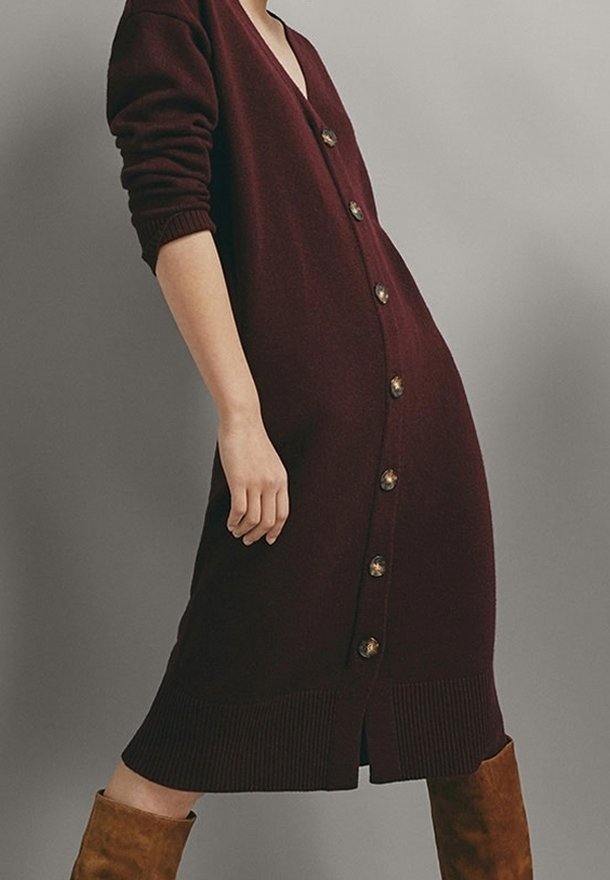 Gros plan d'une personne portant une robe cardigan longue boutonnée de couleur bordeaux foncé et des bottes en daim marron montant jusqu'aux genoux, sur un fond gris uni.