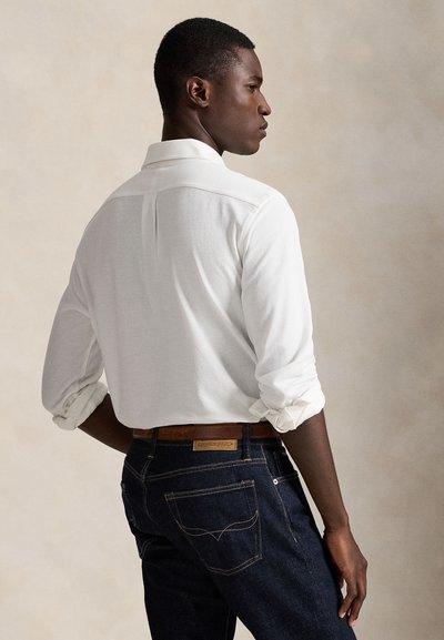 Homme de profil portant une chemise blanche à manches longues avec les poignets retroussés et un jean bleu foncé avec une ceinture marron.