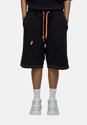Personne portant un short noir avec des cordons et des coutures orange, des chaussettes blanches et des baskets blanches, debout les mains dans les poches.