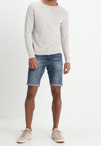 Jasnoszara koszula z długim rękawem, niebieskie szorty dżinsowe z podwiniętymi mankietami oraz beżowe trampki. Tkanina jest gładka, a design casualowy.