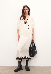 Tejkrém kardigán fekete gombokkal, fehér ruhára rétegezve, amelynek fekete csipke szegélye van. A megjelenést fekete csizmák és egy struktúrált fekete kézitáska teszi teljessé.