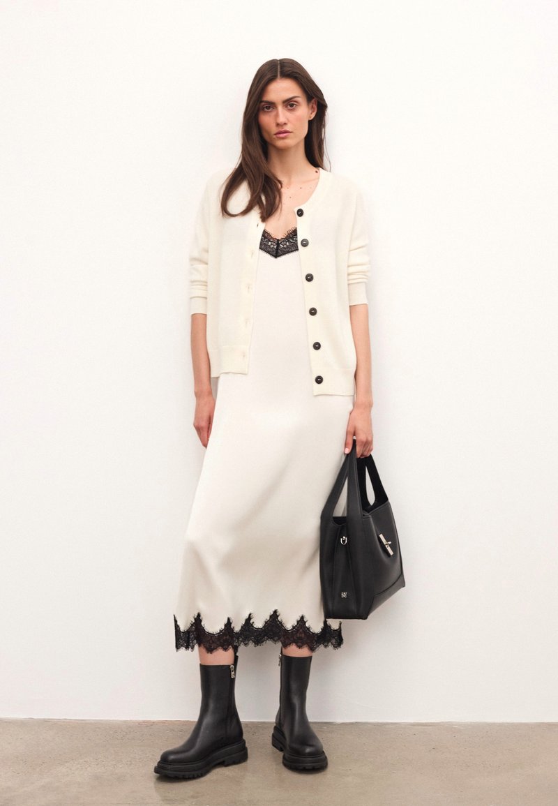 Tejkrém kardigán fekete gombokkal, fehér ruhára rétegezve, amelynek fekete csipke szegélye van. A megjelenést fekete csizmák és egy struktúrált fekete kézitáska teszi teljessé.