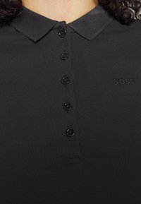 Gros plan d'une chemise noire à col avec cinq boutons et "BOSS" brodé sur la poitrine gauche, portée par une personne aux cheveux bouclés.