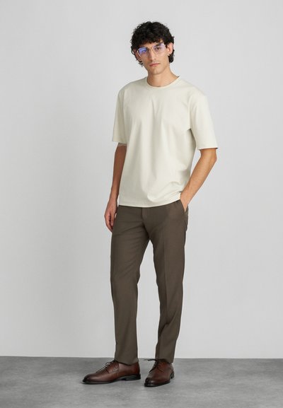 Krém rövidujjú póló, barna slim-fit nadrággal párosítva. A modellen barna alkalmi cipő és átlátszó keretes szemüveg található, világos háttér előtt áll.