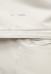 ClimaWarm márkájú, sima textúrájú fehér anyag; varrott varrással és egy kis cipzáras zsebbel az alsó részén.