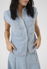 Camicetta in denim azzurro chiaro con maniche corte, apertura frontale con bottoni e due tasche sul petto. Texture morbida, design casual, dettagli di cucitura visibili.