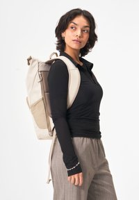 Beige Rucksack mit Mesh-Akzenten und gepolsterten Trägern. Verfügt über ein Roll-Top-Design und mehrere Taschen zur Aufbewahrung. Glatte textile Oberfläche.