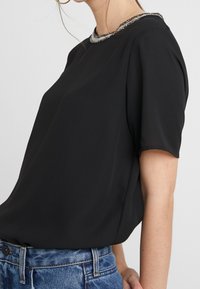 Blusa negra de mangas cortas con un cuello redondo adornado con acentos de cuentas; tejido suave con un corte holgado, combinada con jeans.
