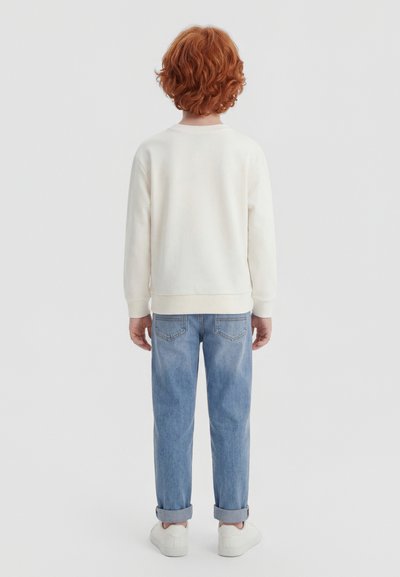Παιδί με σγουρά κόκκινα μαλλιά που φοράει λευκό φούτερ, τζιν μπλε με γυρισμένα μπατζάκια και λευκά αθλητικά παπούτσια, στέκεται γυρισμένο προς τα πίσω πάνω σε απλό φόντο.