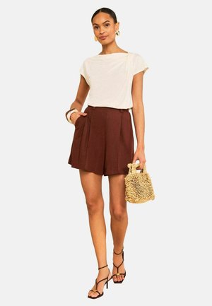 Kvinde iført en hvid kortærmet top, brune højtaljede plisserede shorts, sorte strophæle, holder en beige tekstureret håndtaske.