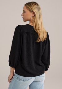 Blusa nera a trama con maniche a sbuffo e motivo a pois. La parte posteriore presenta un giogo, abbinata a jeans in denim chiaro.