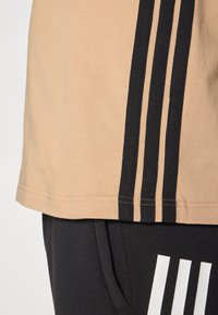 Beige T-shirt med tre lodrette sorte striber på siden, parret med sorte bukser med et hvidt logo. Blødt stof, afslappet pasform.