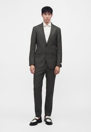 Hombre de pie con traje gris oscuro, camisa blanca y mocasines negros y blancos sobre un fondo blanco liso.