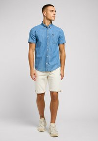 Koszula w krótkim rękawie z denimu w jasnoniebieskim kolorze, z kieszonką na piersi, w zestawieniu z kremowymi szortami. Buty są w jasnym beżu z delikatnymi detalami.