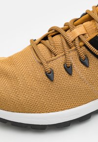 Žlutá strukturovaná sportovní bota s černými šněrovadly a očky, bílým středovým dílem a černou gumovou podrážkou. Detailní pohled.
