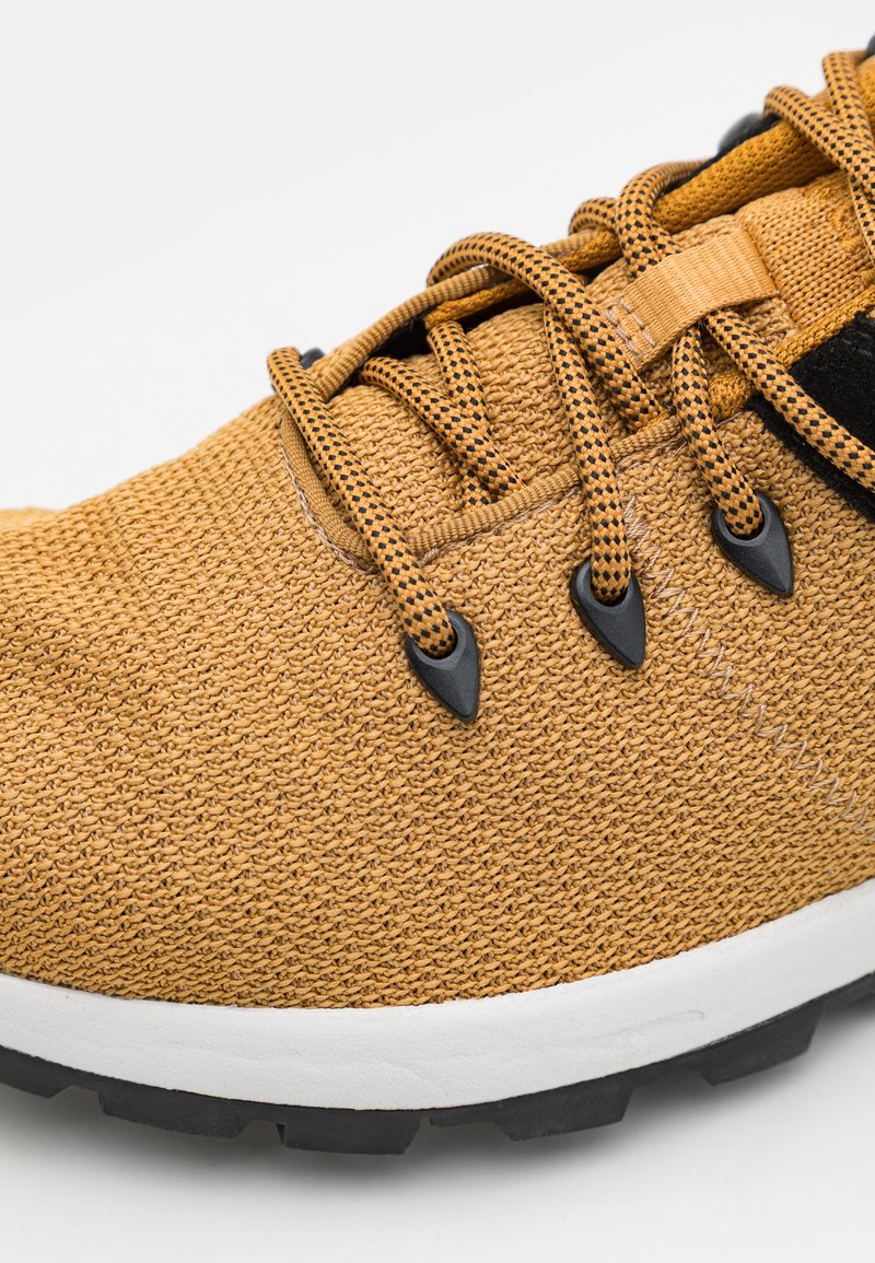 Chaussure de sport texturée jaune avec des lacets et des œillets noirs, une semelle intermédiaire blanche et une semelle extérieure en caoutchouc noir. Vue en gros plan.