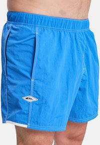 Blue swim shorts made of lightweight fabric. Features an elastic waistband, side pockets, white stitching, and a small logo on the hem.