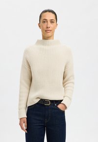 Helles beige, grob gestricktes Sweatshirt mit hohem Kragen, gerippter Textur und langen Ärmeln, kombiniert mit dunkelblauen Jeans und einem schwarzen Gürtel.