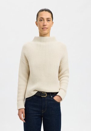 Light beige chunky knit sweater with a high neck, ribbed texture, and long sleeves, paired with dark blue jeans and a black belt.
